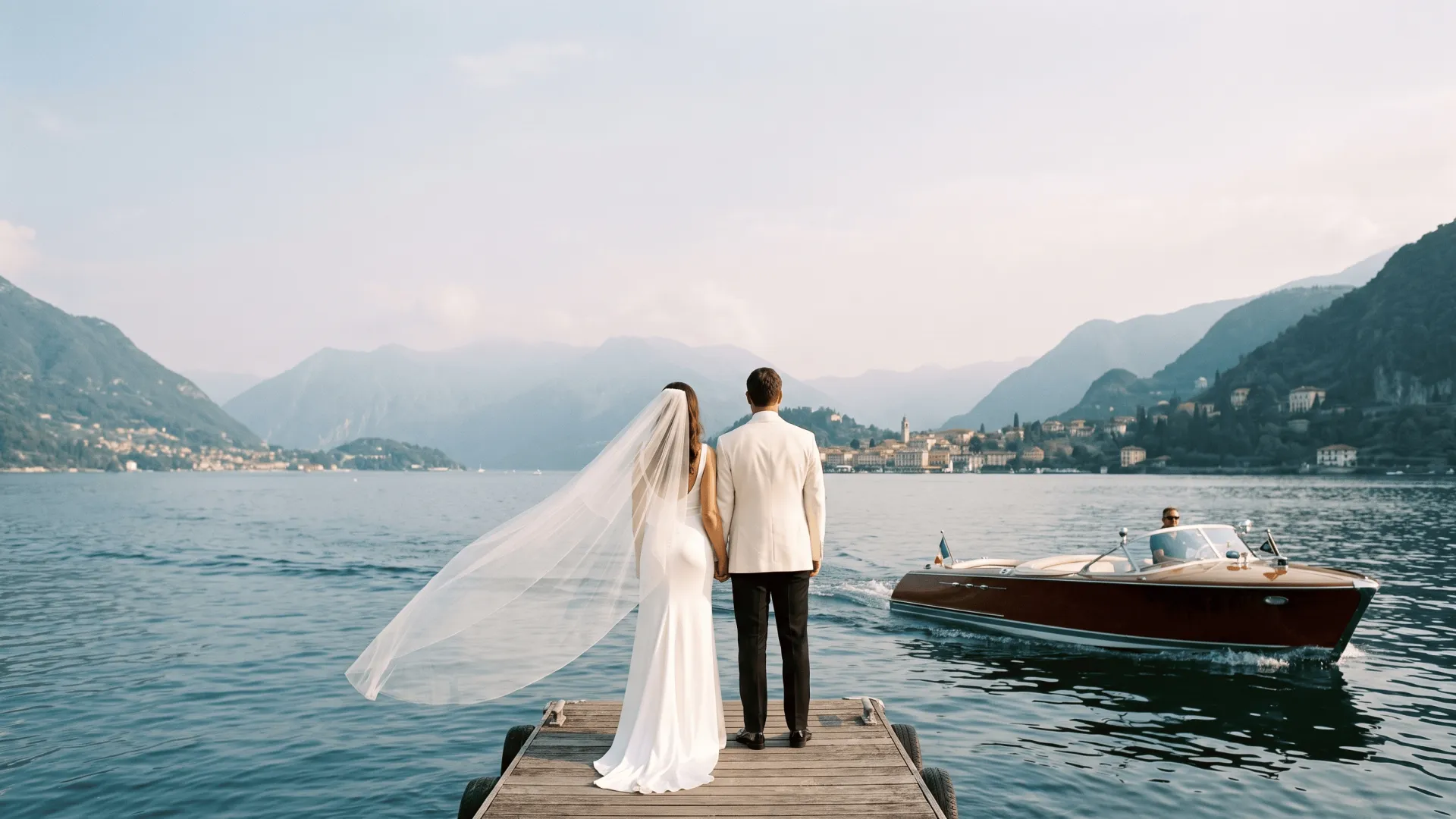 A couple viewed from behind beside a European lake as a wooden boat passes